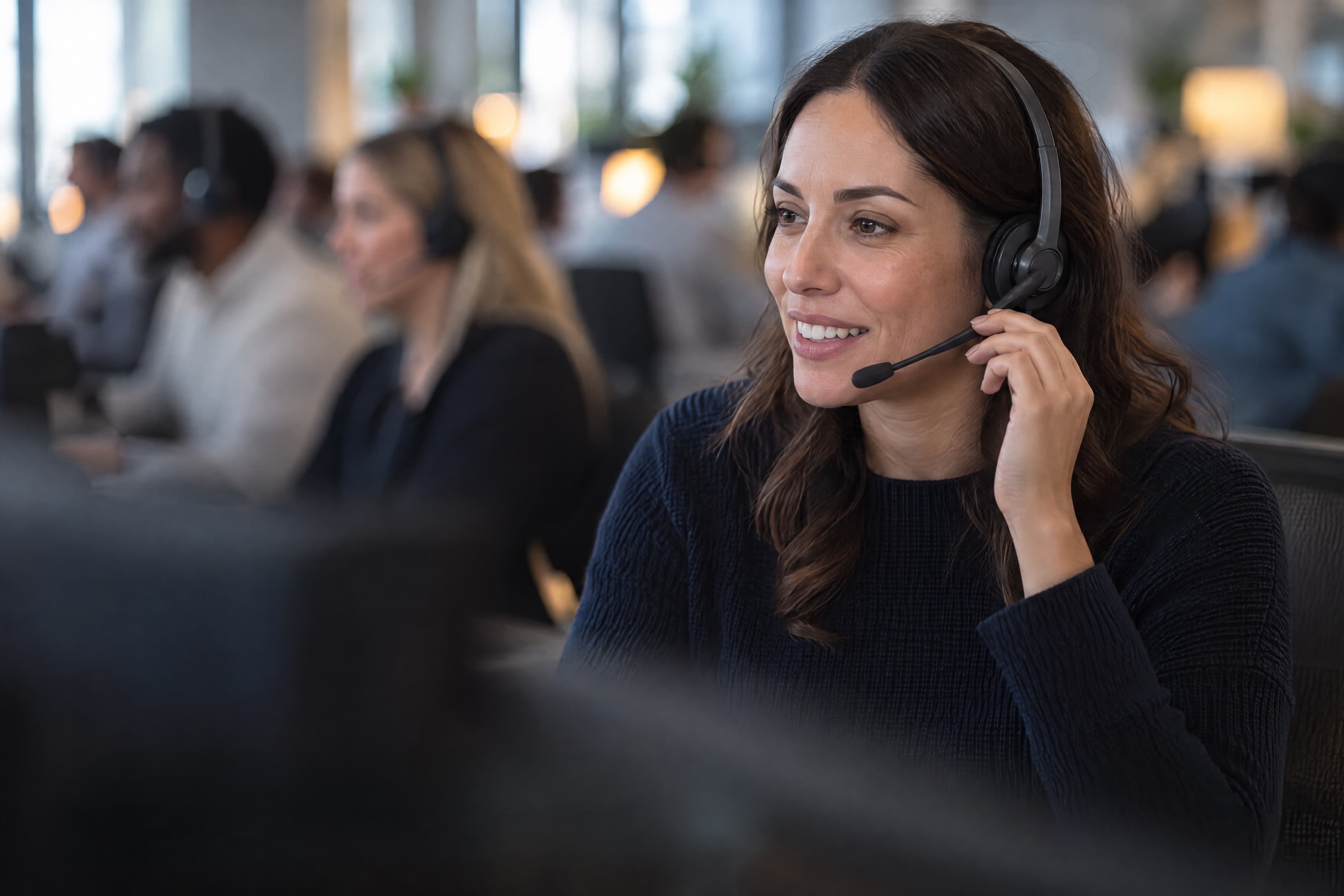 Un’operatrice di call center con le cuffie, alle sue spalle colleghi che parlano alle loro postazioni.